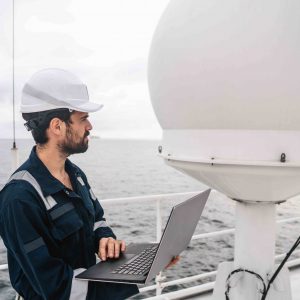 Marine service technician or serviceman near VSAT terminal on deck of vessel or ship. He is working on laptop or notebook